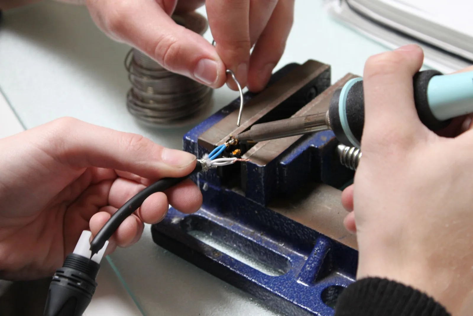 Photo of an XLR insert clamped in a vise with the cable housing already threaded on, showing solder being fed into a heated terminal joint.